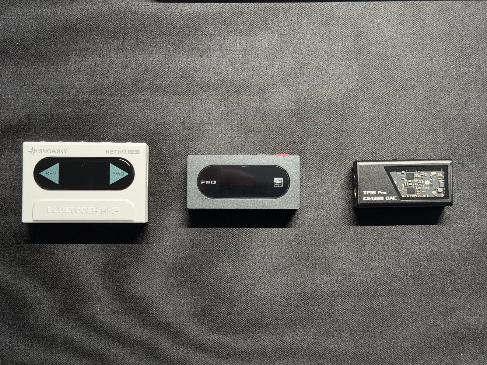 Ba dongle DAC/amp nhỏ đặt cạnh nhau trên nền vải tối — mỗi cái một kiểu dáng